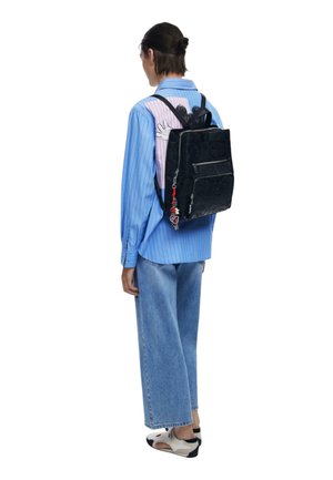 Person wearing a blue striped shirt with cartoon graphics, wide-leg jeans, white sneakers, and a black embossed backpack with keychain accessories.