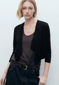 Svart kort cardigan med öppen front och långa ärmar, bärs över en brun topp och mörk denimjeans, med en mjuk yta och minimalistisk design.
