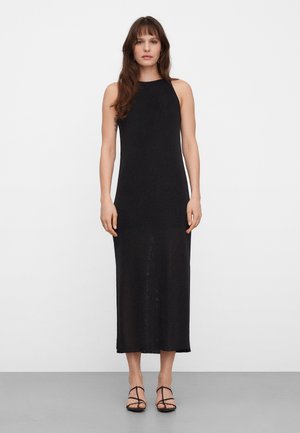 Vrouw staand tegen een witte achtergrond, gekleed in een mouwloze zwarte midi-jurk en zwarte sandalen met hak en bandjes.