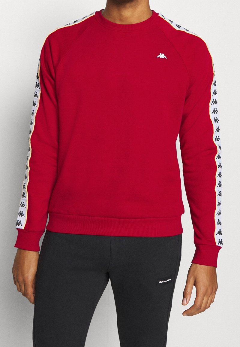 Homme portant un sweat-shirt rouge avec un ruban logo blanc et noir sur les manches, associé à un pantalon de jogging noir avec un petit logo blanc.