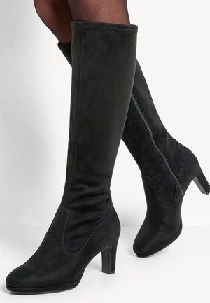 Bottes noires en daim montant jusqu'aux genoux avec talons carrés et fermeture éclair latérale, portées avec des collants foncés sur fond blanc.