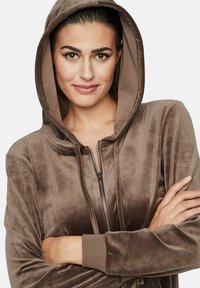 Hoodie en velours marron avec zip sur le devant, poches latérales et poignets côtelés. Le tissu présente une finition douce et texturée.
