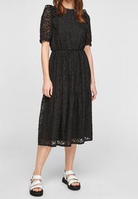 Robe midi en dentelle noire avec de courtes manches bouffantes, taille élastique, jupe à volants, motif floral et accents à volants. Assortie avec des sandales blanches.