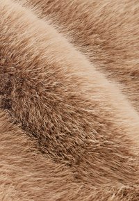 Tissu en fausse fourrure beige avec des fibres douces et longues, présentant un léger brillant, une texture rugueuse et des variations subtiles de couleur.