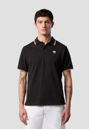 Giovane uomo con capelli corti e scuri che indossa una polo nera con rifiniture arancioni e pantaloni bianchi, in piedi davanti a uno sfondo grigio semplice.