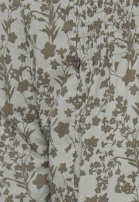 Lys stof med et grønt blomstermønster, der har mørkegrønne blomster og blade. Inkluderer smock-detalje øverst og en lille metallisk accent.