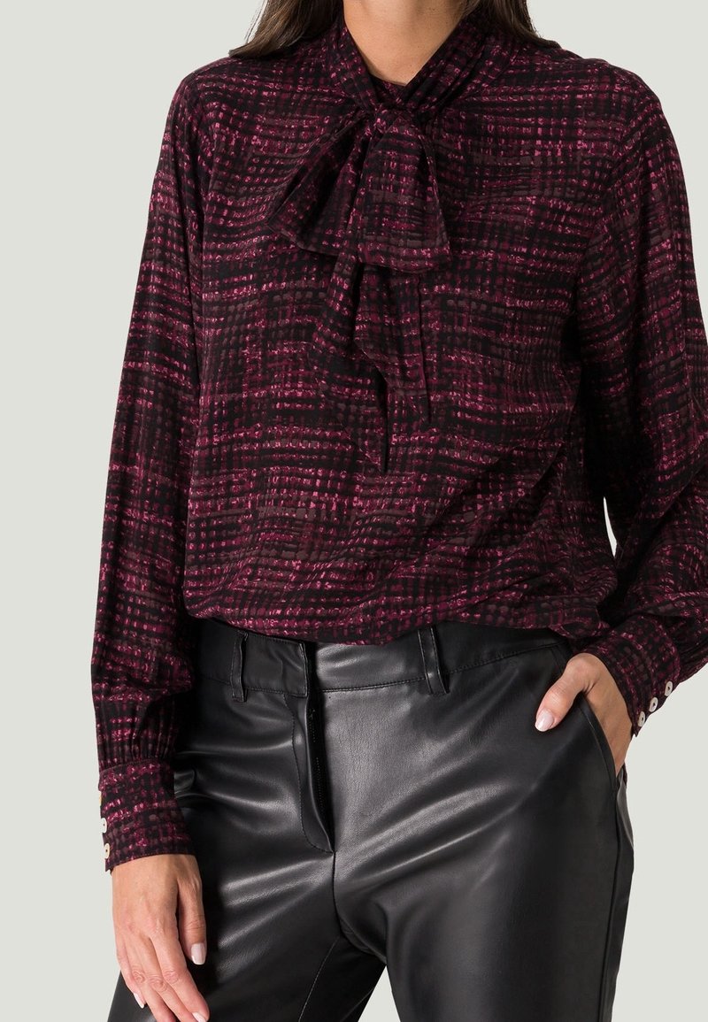 Blouse à manches longues de couleur bordeaux foncé avec un motif géométrique ; elle présente un col à nœud papillon et des poignets boutonnés, associée à un pantalon en cuir noir.