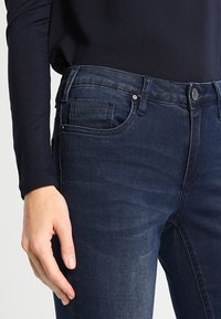Dark blue denim jeans with a slim fit, five-pocket design, light fading, and a button closure at the waist. Worn with a long-sleeve top.