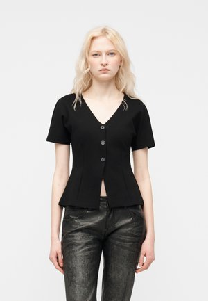 Ξανθιά γυναίκα που φοράει μαύρο εφαρμοστό πουκάμισο με κοντά μανίκια, V-λαιμόκοψη και κουμπιά, καθώς και μαύρο δερμάτινο παντελόνι με υφή, στέκεται μπροστά σε απλό φόντο.