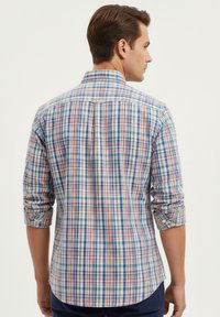 Hombre de espaldas vistiendo una camisa de cuadros multicolor de manga larga con las mangas arremangadas y pantalones oscuros, contra un fondo liso.