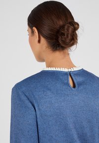 Blauwe trui met een gestructureerde afwerking, voorzien van een witte gerimpelde kraag en een keyhole-detail aan de achterkant, vastgezet met een knoop.