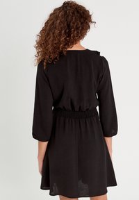 Femme aux cheveux bouclés portant une robe noire à manches longues avec une taille élastique, se tenant de dos contre un fond uni.