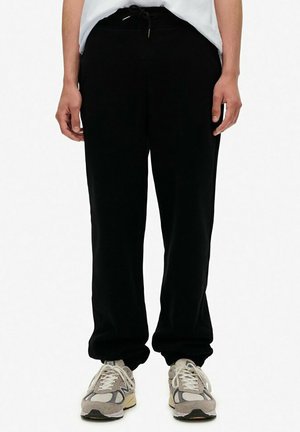 Person trägt schwarze Sweatpants mit Kordelzug am Bund und elastischen Bündchen, kombiniert mit weißen und grauen Sneakers, stehend vor weißem Hintergrund.