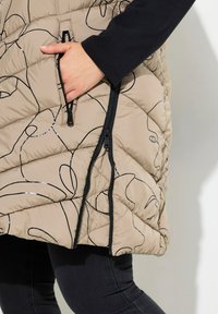 Beige gesteppte Pufferweste mit schwarzem Linienstil, ausgestattet mit einer Reißverschlusstasche und kontrastierendem Reißverschluss, kombiniert mit dunklen Jeans.