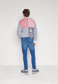 Pull avec un design coloré en blocs comprenant des teintes de rose, gris et blanc ; porté avec un jean bleu et des chaussures de sport grises.