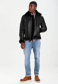 Chaqueta bomber de cuero negro con un cuello y puños de pelaje sintético, combinada con una camisa de cuadros, jeans de mezclilla azul claro y botas marrones.