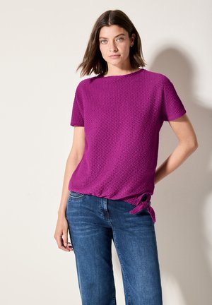 Top de manga corta, texturizado en un púrpura vibrante con cuello redondo y detalle atado en el dobladillo, combinado con jeans de mezclilla azules.