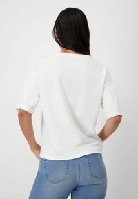 Bílý triko s krátkým rukávem, volného střihu a kulatým výstřihem. Pozadí je čistě bílé, což zdůrazňuje design oděvu.