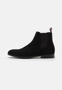 Bottines Chelsea en daim noir avec panneaux latéraux élastiques, bout arrondi, talon bas et languette rouge à l'arrière. Fini lisse et texturé.