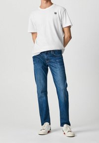 Vit bomull t-shirt med en svart logotyp på vänster bröst, tillsammans med blå denimjeans. Skorna är vita sneakers med accenter.