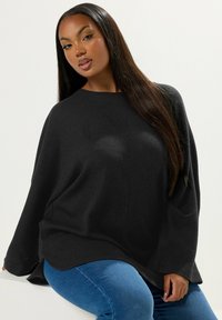Schwarzer gerippter Pullover mit lockerer Passform, weiten Ärmeln und rundem Ausschnitt, kombiniert mit blauen Jeans. Glatte Textur, minimale Details.