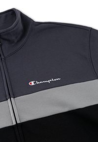 Felpa con zip grigia scura e colletto alto, caratterizzata da una striscia grigia orizzontale e una parte inferiore nera. Logo "Champion" in bianco.