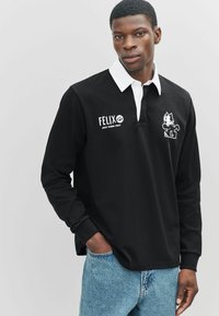 Polo negro de manga larga con cuello blanco, con bordado de Felix el Gato y texto en el lado izquierdo. Llevado con vaqueros azules.