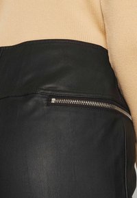 Zwarte leren rok met een ritssluiting zak. Heeft een gladde textuur en afgestikte naden, gecombineerd met een beige gebreid topje dat zichtbaar is erboven.