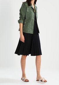 Femme portant une veste vert olive sur une robe noire plissée jusqu'aux genoux et des sandales plates beiges sur fond blanc.