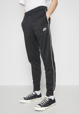 Osoba ubrana w czarne spodnie dresowe Nike z białymi paskami po bokach oraz czarne niskie sneakersy z białymi sznurowadłami i podeszwami. Ręce w kieszeniach.