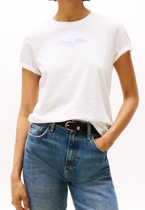 Persona que lleva una camiseta blanca de Tommy Hilfiger metida en unos vaqueros azules con un cinturón negro, una mano en el bolsillo, sobre un fondo blanco.