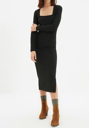 Femme portant une robe midi noire à manches longues, des chaussettes grises et des bottines en daim marron, debout avec un bras croisé sur l’autre.