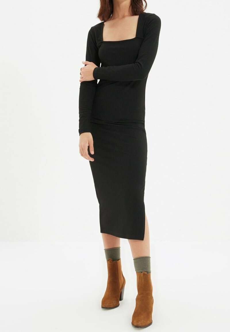 Femme portant une robe midi noire à manches longues, des chaussettes grises et des bottines en daim marron, debout avec un bras croisé sur l’autre.
