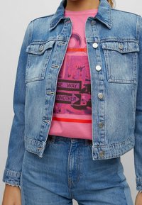 Osoba oblečená v světle modré džínsové bundě přes růžové tričko s grafickým potiskem, na kterém je text "ONE WAY" a "ANOTHER", doplněná modrými džínsami.