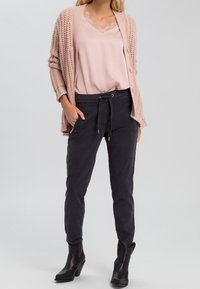 Cardigan rosa chiaro lavorato a maglia sopra una maglietta rosa con pizzo, abbinato a pantaloni grigi scuri con cordoncino e tasche con zip. Stivali neri alla caviglia.