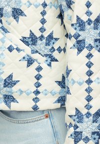 Κοντινό πλάνο επενδύτη με σχέδιο από μπλε αστέρια σε πατς, φορεμένο πάνω από ανοιχτόχρωμο τζιν παντελόνι.