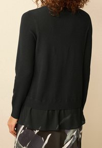 Gestreifter schwarzer Pullover mit langen Ärmeln und einem geschichteten Rüschensaum. Glatte Textur, figurbetontes Design und subtile Variationen in der Stoffdicke.