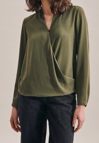 Blusa verde oliva a maniche lunghe con scollatura a V, dettaglio drappeggiato sul davanti e accentuazione con bottone. Abbinata a jeans in denim scuro. Tessuto dalla texture liscia.