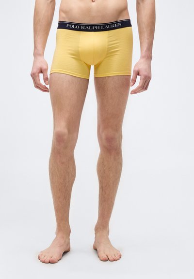Ανδρικό σώμα και πόδια που φορούν κίτρινα εσώρουχα Polo Ralph Lauren με μαύρη μέση, όρθιος ξυπόλητος σε απλό λευκό φόντο.
