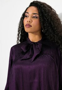 Blusa viola con una finitura texturizzata, caratterizzata da un collo a fiocco prominente e maniche lunghe. Il tessuto ha una lucentezza satinata e strisce verticali.