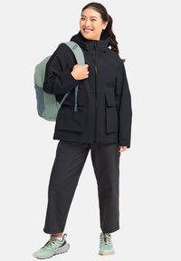 Giacca impermeabile nera con due tasche frontali, colletto alto e cappuccio. Abbinata a pantaloni scuri e sneakers color verde acqua chiaro. Zaino verde chiaro.