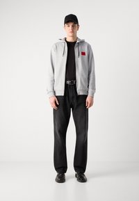 Šedá mikina na zip s červeným logem, černé tričko, tmavé džíny a černé tenisky. Model stojí s rukama po stranách na neutrálním pozadí.