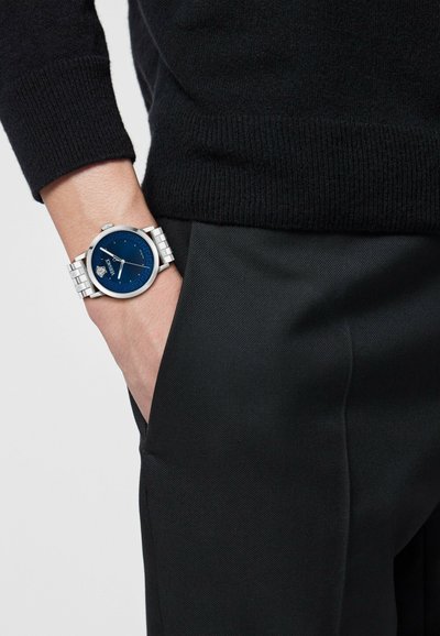 Srebrny zegarek na nadgarstek z okrągłą niebieską tarczą, metalową bransoletką i białymi znacznikami godzin. Noszony na nadgarstku z czarnym swetrem z dzianiny.
