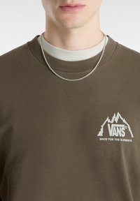 Bruine sweatshirt met geribbelde halslijn, met een witte berglogo en de tekst "VANS MADE FOR THE ELEMENTS" op de borst. Zilveren ketting.