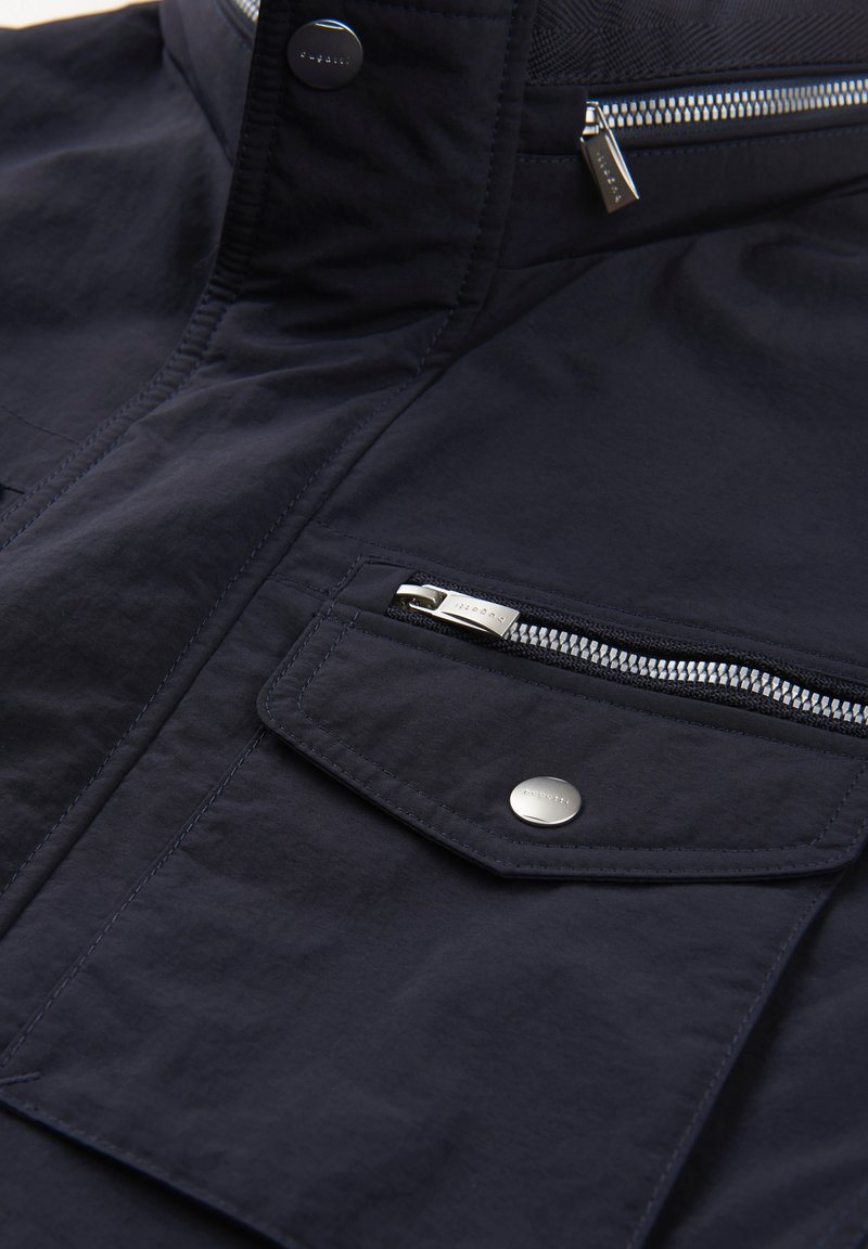Marineblaue Jacke mit glatter Textur, Vorderknopf, Reißverschlusstasche und gestickten Details. Funktionales Design mit modernen Akzenten.