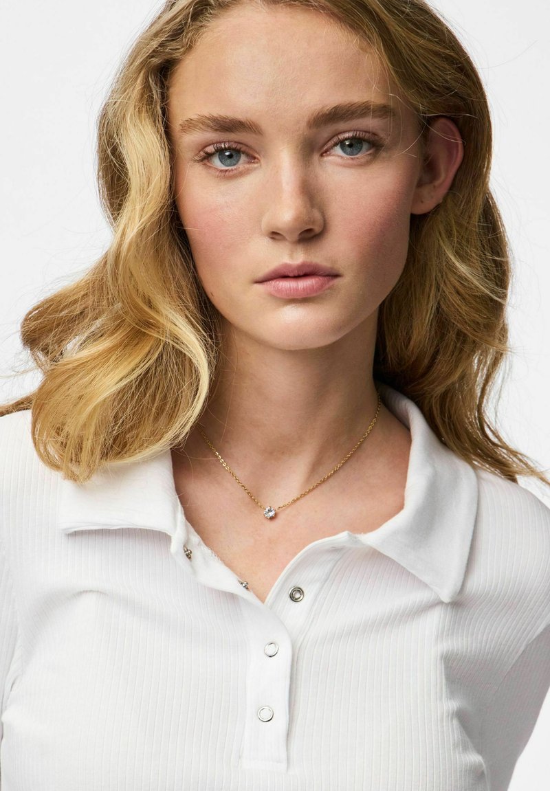 Polo blanc côtelé avec col et patte de boutonnage ; présente un délicat collier en or avec une pierre centrale. Texture douce et design classique.