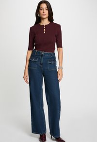 Frau, die ein burgunderrot gestricktes Oberteil mit Knopfdetails und weit geschnittenen blauen Jeans mit vorderen Klappentaschen trägt, steht vor einem einfarbigen Hintergrund.