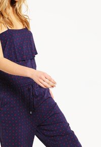 Marinblå jumpsuit med rynkad topp och röd blommönster. Har ett elastiskt midjeband och justerbar dragsko. Lättviktig tyg.