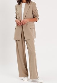 Blazer beige ajusté avec des revers et deux poches avant, associé à un pantalon ample assorti. Porté avec un haut blanc et des baskets blanches.