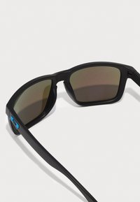 Schwarze Sonnenbrille mit rechteckigen Rahmen, polarisierten braunen Gläsern, strukturiertem schwarzen Kunststoff und einem kontrastierenden blauen Akzent an der Seite.